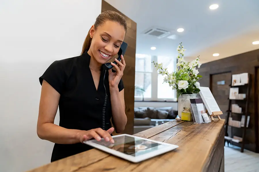 A smiling aesthetics clinic receptionist wearing a black tunic speaks to a customer on the phone and logs a new appointment on a tablet.